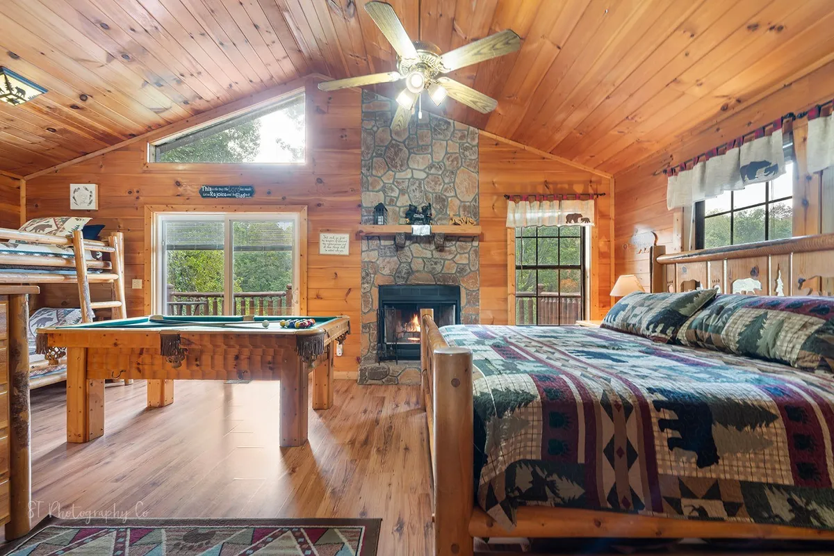 A cozy view of the suite with the stone fireplace glowing warmly beside the log bed, twin bunk, and pool table. The scene radiates togetherness, with ample space for lounging, storytelling, and friendly game tournaments.