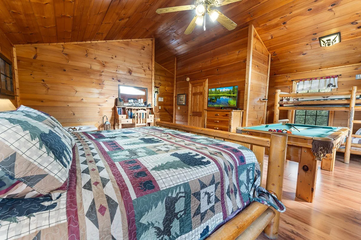 This inviting upstairs suite features a king log bed, cozy twin bunk beds, a pool table at the center, and a TV, all set against rustic wooden walls. The warm lighting and soft textiles enhance the room’s welcoming, family-friendly feel, while the pool table signals playful fun within the sleeping quarters.