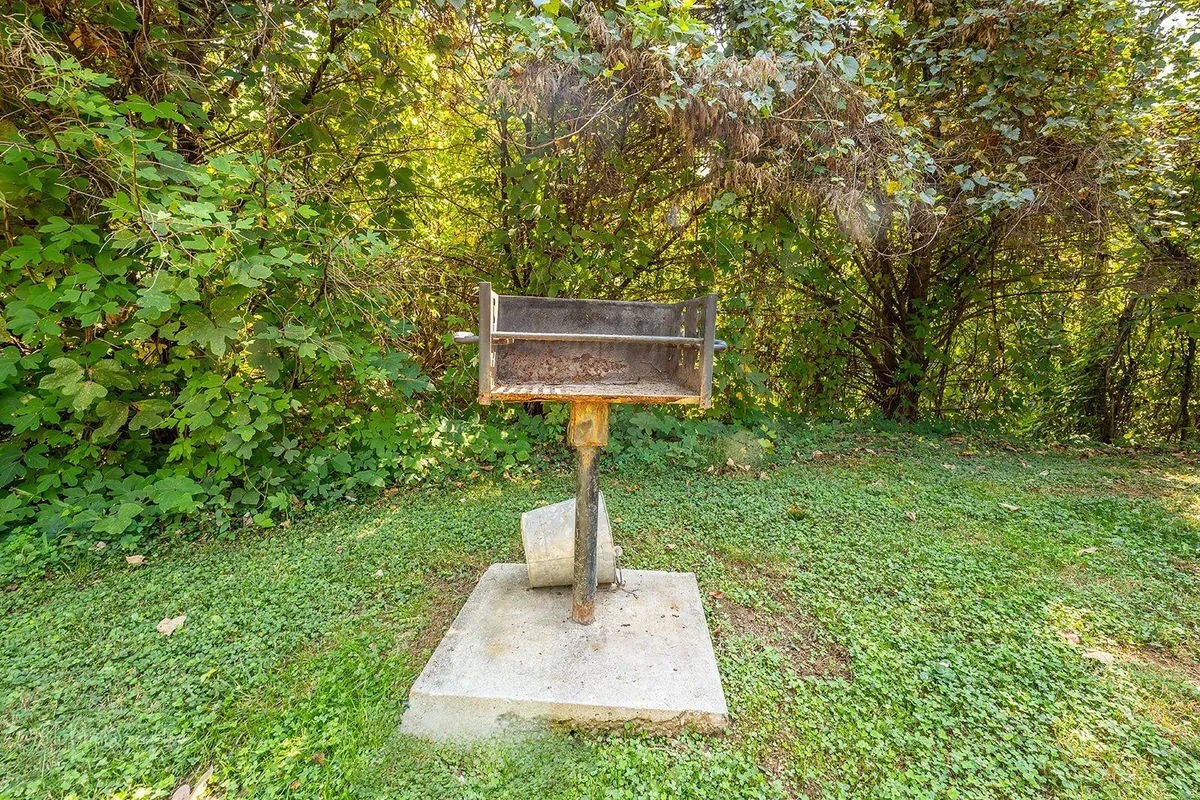 A dedicated charcoal grill stands to one side, framed by lush greenery, signaling a space meant for cookouts and communal meals. The setup is simple and inviting, encouraging barbecuing and gathering outdoors.