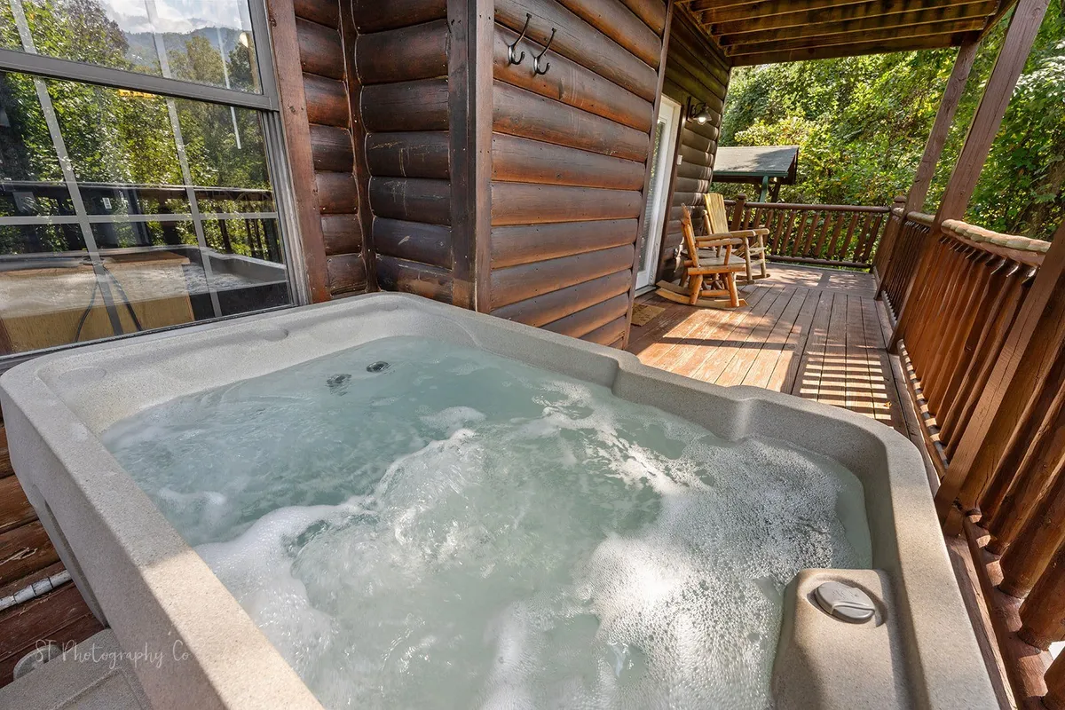 A tranquil scene featuring a bubbling hot tub on a wooden deck, surrounded by lush greenery. Two rocking chairs are nearby, inviting relaxation while taking in the fresh mountain air and scenic views. The warm, inviting water and open-air setting evoke a sense of privacy and luxury.
