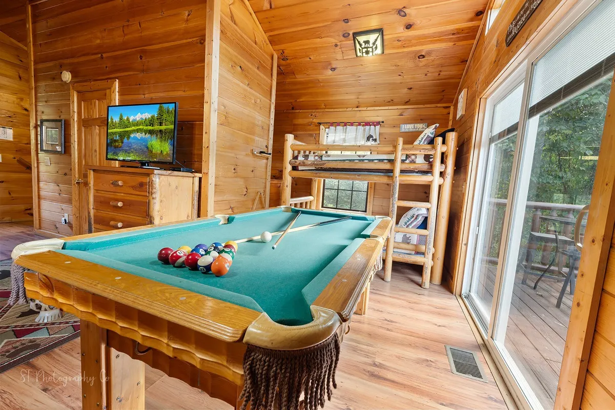 A spacious family game and relaxation area features a pool table, bunk beds, a wall-mounted TV, and a sliding glass door that opens to sunset views. The room buzzes with activity, from friendly pool competitions to movie nights and quiet time on the bunks.