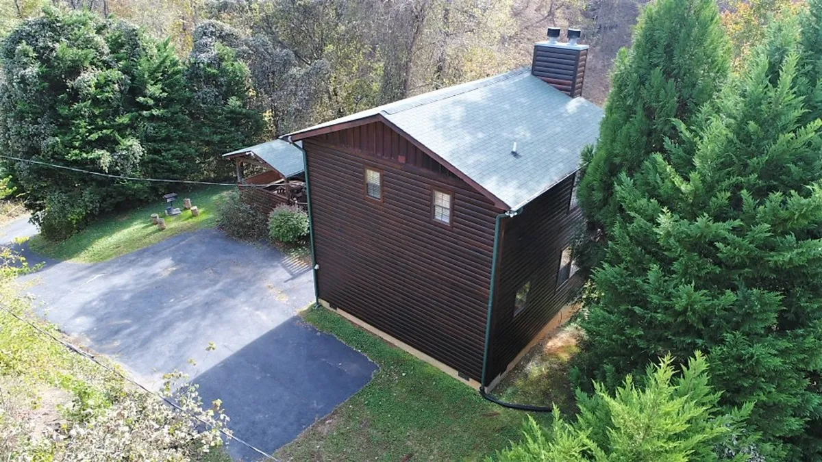 This exterior shot showcases the cabin's wooden siding nestled among mature trees, with a driveway leading up and a sense of privacy provided by the foliage. Natural light filters through the branches, creating a welcoming and secluded atmosphere.