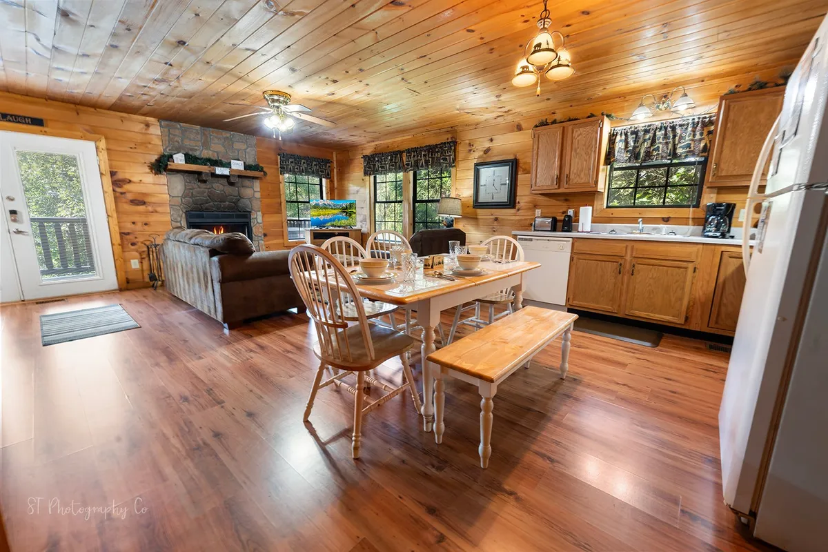 This image showcases an open-concept living and dining area, with a rustic wooden dining table set for six, a cozy stone fireplace, comfortable seating, large windows with nature views, and a kitchen area visible in the background. The warm, homely atmosphere is highlighted by the soft lighting and natural textures.