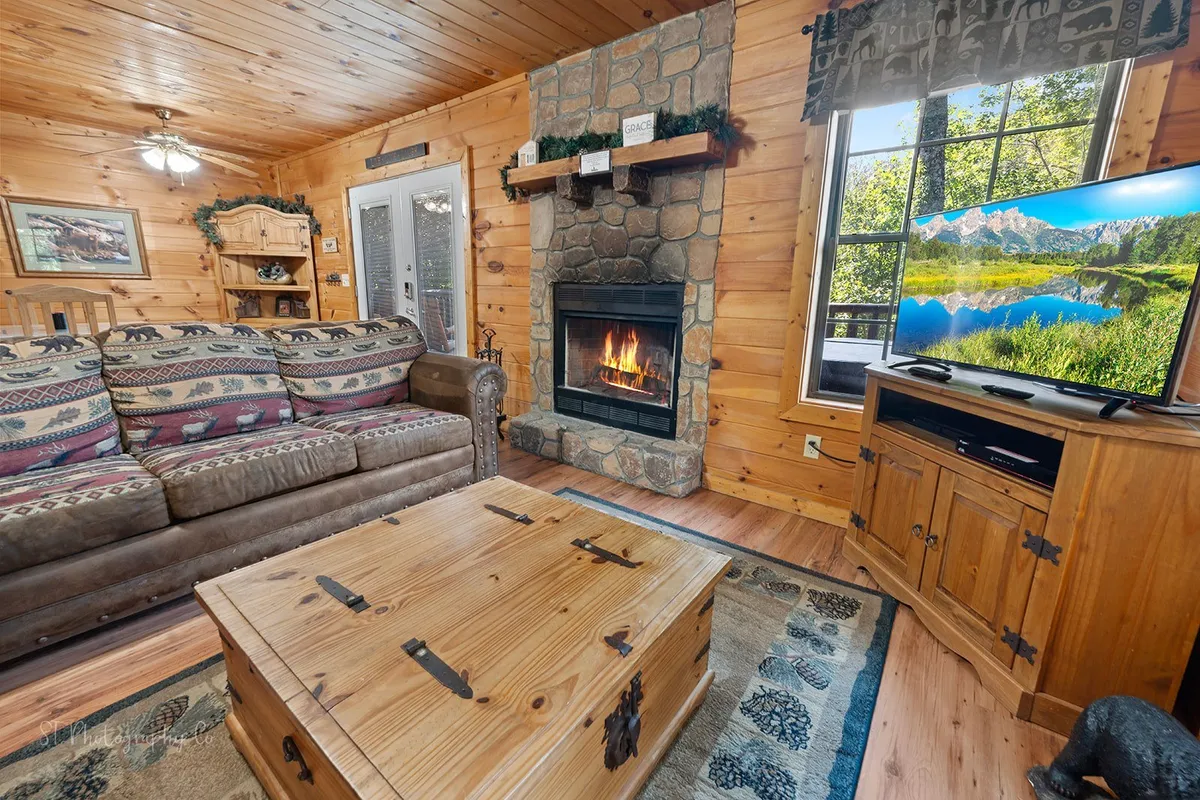 The image features a cozy living area with a welcoming sofa arranged around a stone fireplace. A coffee table sits in front, and a television is mounted nearby, framed by large windows that let in natural light and offer glimpses of the outside scenery. The decor is warm and rustic, evoking a sense of relaxation and comfort.
