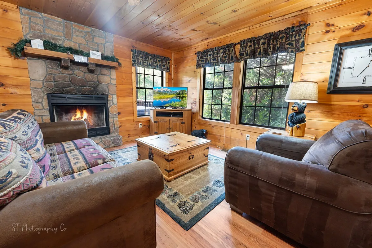 A welcoming living room scene featuring a prominent stone fireplace, wooden furniture, textured cushions, and expansive windows that let in natural light. The furnishings invite relaxation and socialization, while the fireplace forms a cozy focal point.