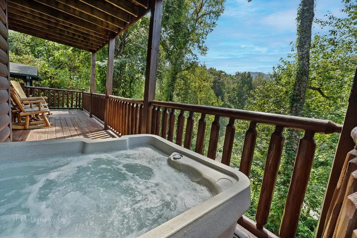 This image captures a tranquil exterior deck scene featuring a bubbling hot tub, wooden railing, rocking chairs, and lush greenery in the background. The space is designed for both relaxation in the hot tub and peaceful contemplation in the rocking chairs, all set against a natural wooded backdrop.